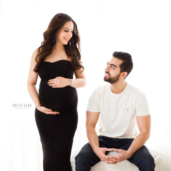 Expecting mother in a black strapless fitted gown smiling at her seated husband in a white tee and navy trousers, bright white backdrop