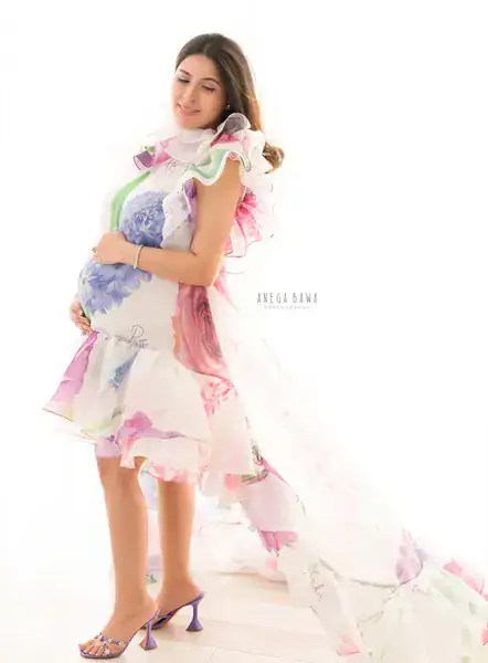 Expecting mother in a floral-print organza mini dress with ruffles and a long sheer floral cape, holding her bump, eyes closed, coloured