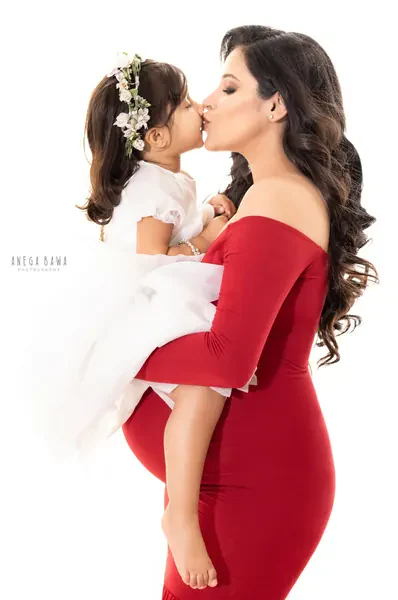 Expecting mother in a red off-shoulder long-sleeve fitted gown holding her young daughter in a white tulle dress with a flower crown