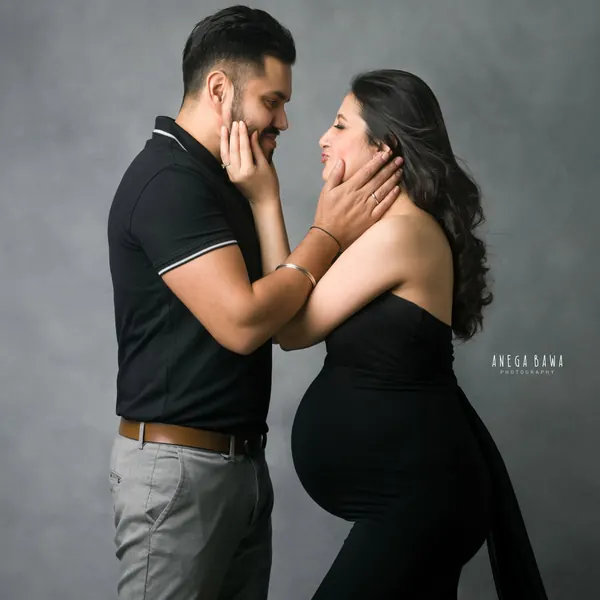 Couple facing each other, husband in a black polo and grey trousers holding his wife's face, she wears a black strapless gown with a draped