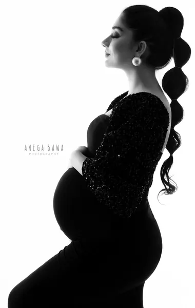 High-key black and white silhouette of expecting mother in profile in a black gown with a sequin sleeve and a long bubble ponytail with