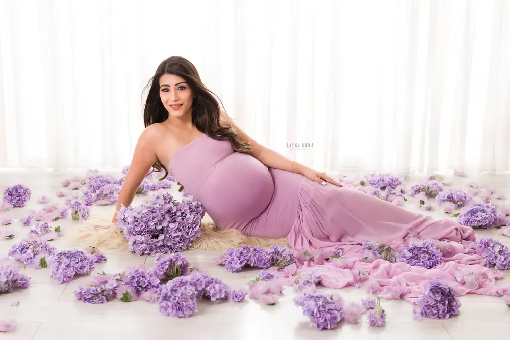 Expecting mother reclining in a lilac strapless fitted gown among purple hydrangeas with a long train on a white floor, sheer curtain