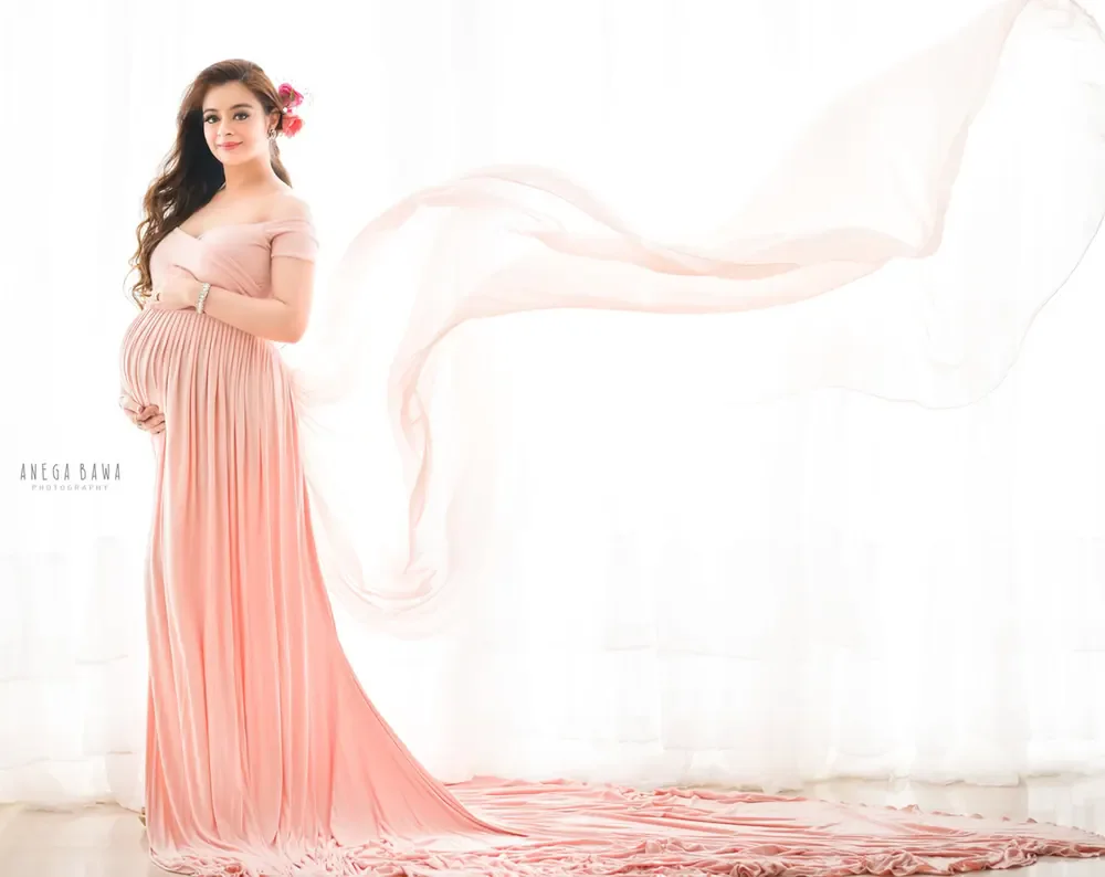 Expecting mother in a peach pink off-shoulder pleated gown with a long train and sheer fabric flying out to the side, a flower in her hair
