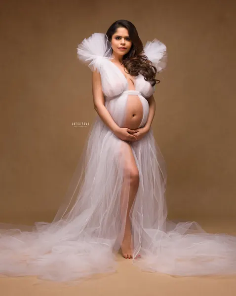 Expecting mother in a white tulle gown with puffed shoulders, open front, high slit and a long tulle train, barefoot, hands cradling her