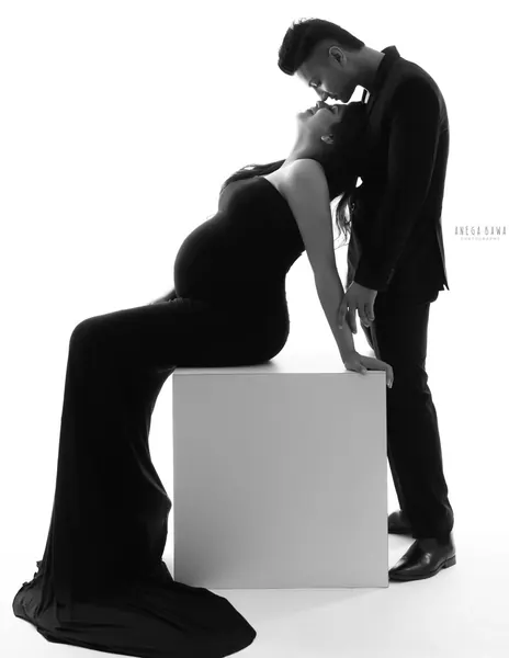 High-key black silhouette of a couple, wife leaning back seated on a white block in a black fitted gown with a long train while her husband