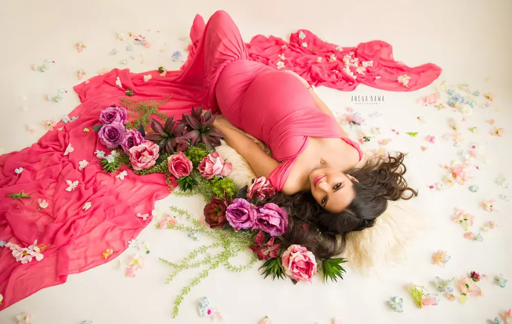 Overhead view of expecting mother lying on a fur rug in a hot pink gown with a long pink train, surrounded by roses and scattered petals on