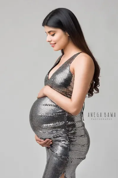 Smiling expecting mother in profile in a silver sequin halter midi dress with a plunging neckline, looking down at her bump, light grey