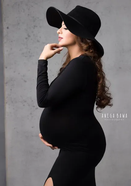 Expecting mother in profile in a fitted black long-sleeve dress with a side slit and a wide-brim black hat, hand to her chin, long curled