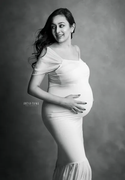 Black and white portrait of smiling expecting mother in a white fitted gown with draped sleeves and a flared hem, looking over her
