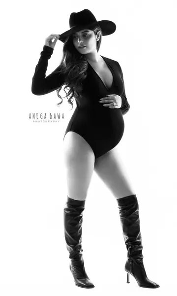 High-key black and white portrait of expecting mother in a black long-sleeve plunge bodysuit, wide-brim hat and knee-high boots, hand on
