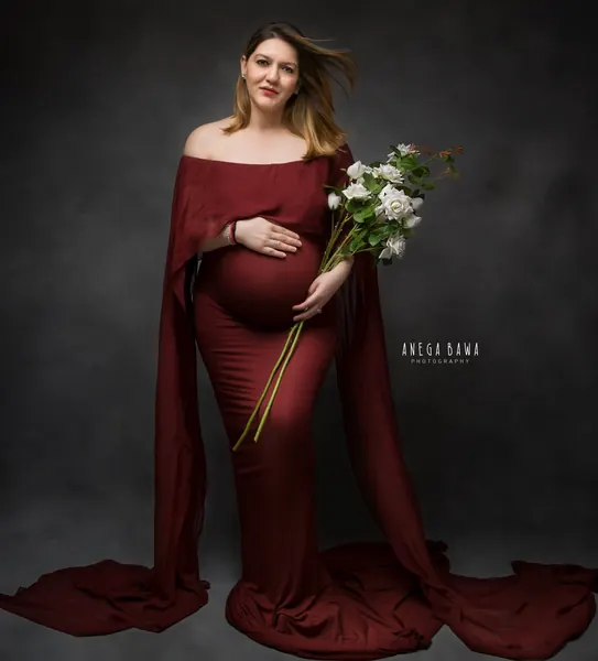 Expecting mother in a maroon off-shoulder gown with long cape sleeves and a train, holding a bunch of white roses, hair blowing, dark grey
