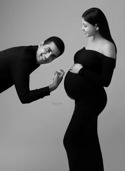 Black and white playful couple portrait: husband in a black shirt leaning in with a surprised smile toward the bump, wife in a black