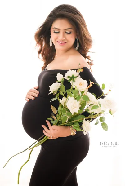 Smiling expecting mother in a black off-shoulder long-sleeve fitted gown with drop earrings holding a bunch of white roses, bright white