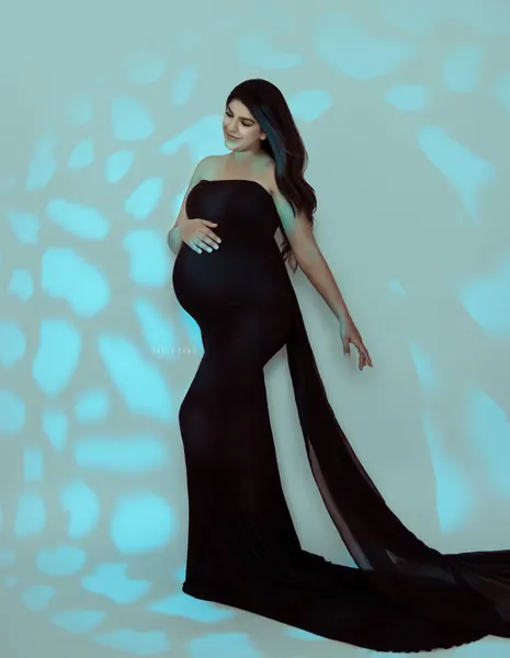 Expecting mother in a fitted black strapless gown with a sheer black train, hand on her bump, turquoise backdrop with patterned light