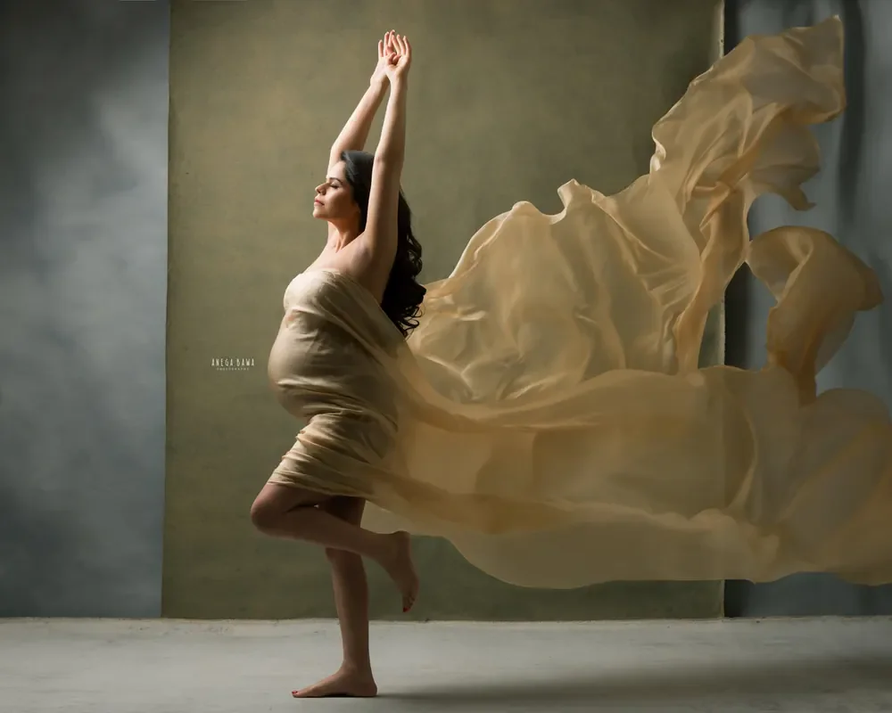 Expecting mother dancing in a champagne silk drape on one leg with arms raised and fabric flying out to the side, barefoot, olive and grey