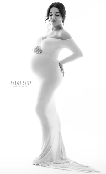High-key black and white portrait of expecting mother in a white off-shoulder fitted gown with a train, hand on her bump and one on her