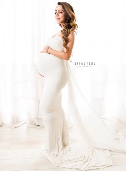 Expecting mother in profile in an ivory white strapless fitted gown with a long draped train, wavy hair, looking down at her bump, bright