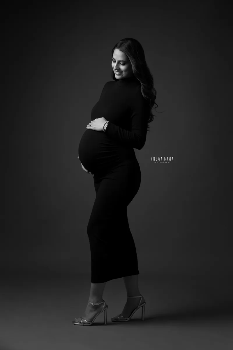 Maternity photography in Delhi: black and white full-length portrait of smiling expecting mother in a fitted black high-neck dress and