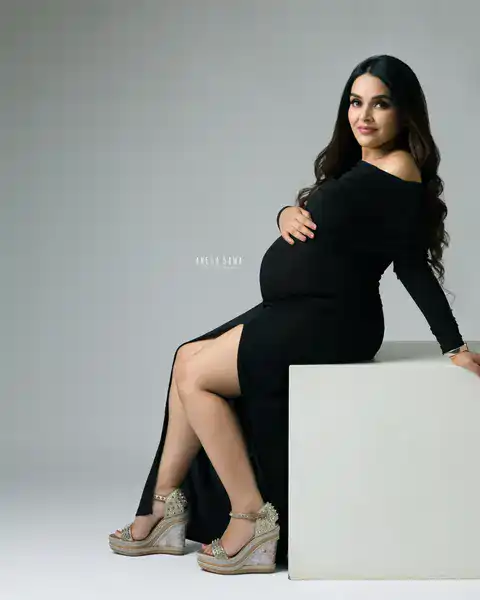 Maternity photography in Delhi: expecting mother seated on a white block in a black off-shoulder gown with a thigh-high slit and
