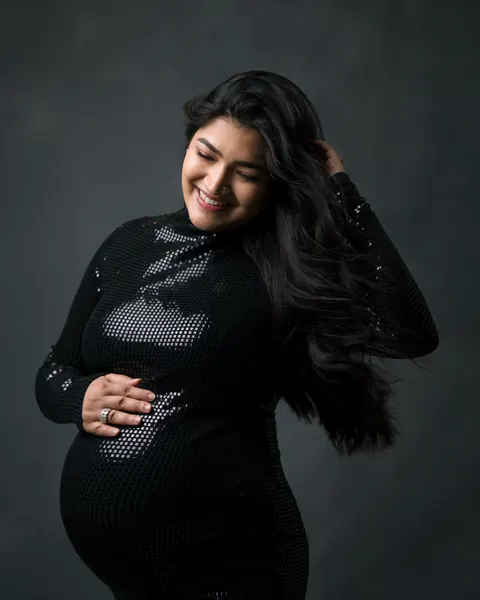 Maternity photography in Delhi: smiling expecting mother with long black hair in a fitted black sequin dress, one hand in her hair and one