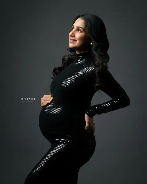 Maternity photography in Delhi: smiling expecting mother in profile in a fitted high-neck black sequin gown, looking upward with a hand on