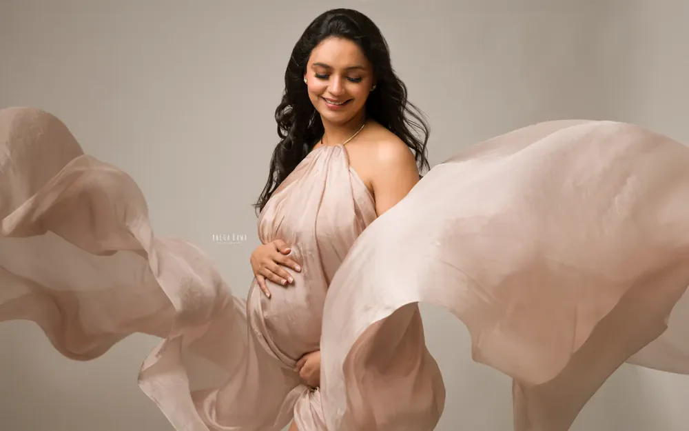 Maternity photography in Delhi: expecting mother in a blush silk halter drape with the fabric flowing out around her, smiling down at her