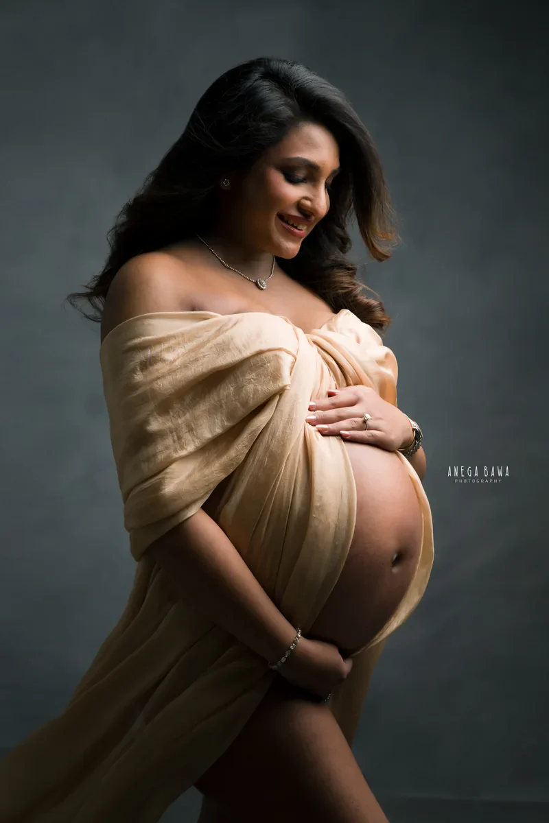 Maternity photography in Delhi: smiling expecting mother in a champagne silk drape off the shoulders, looking down and holding her bump