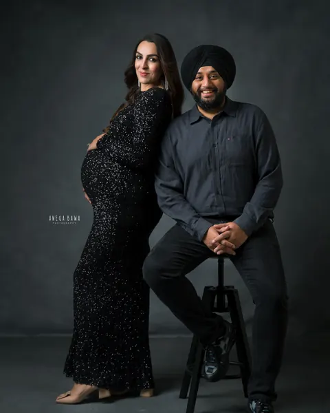 Maternity photography in Delhi: smiling couple, wife in a black sequin gown leaning back and husband in a black turban seated on a stool