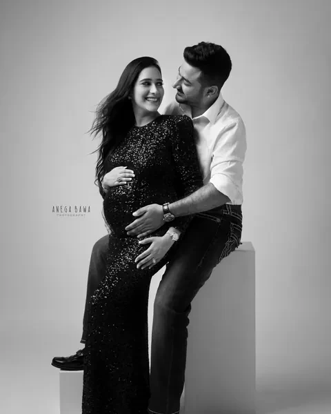 Maternity photography in Delhi: black and white portrait of a laughing couple, husband seated on a block hugging his wife in a sequin gown