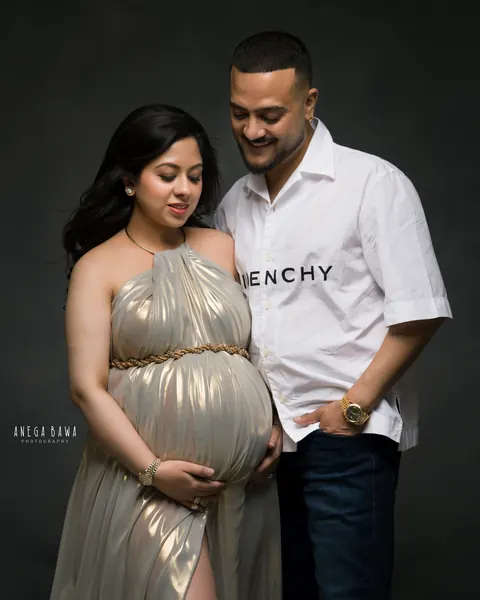 Maternity photography in Delhi: couple looking down at the bump, wife in a gold metallic halter gown with a rope belt and a side slit