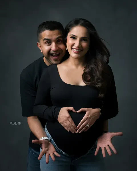 Maternity photography in Delhi: playful couple, wife making a heart shape with her hands on her bump while her husband peeks over her
