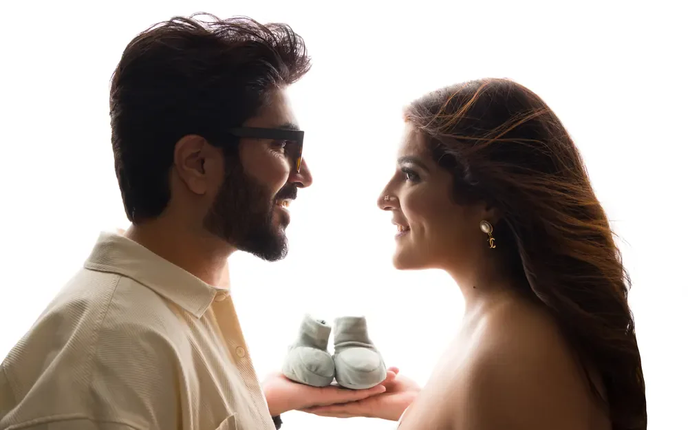 Maternity photography in Delhi: couple face to face in profile holding a pair of baby booties between their hands, bright white backdrop