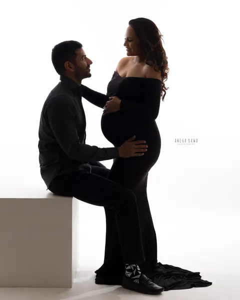 Maternity photography in Delhi: backlit silhouette of a husband seated on a white block with his hand on the bump of his wife, who wears an