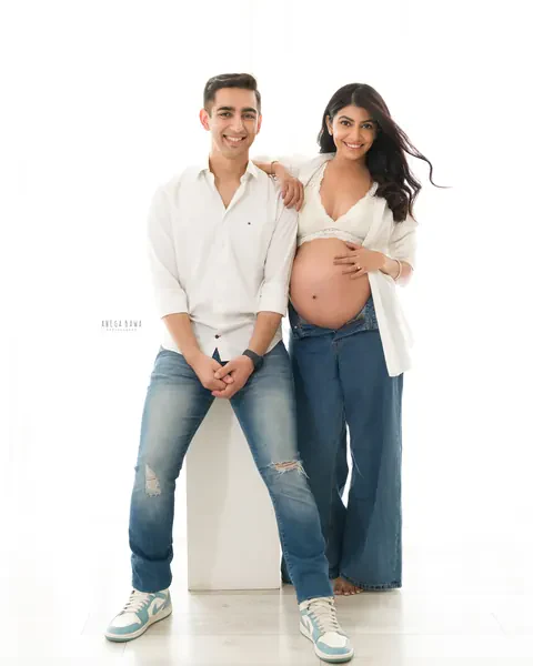 Maternity photography in Delhi: smiling couple, husband seated on a white block in a white shirt and jeans and wife leaning on him in a