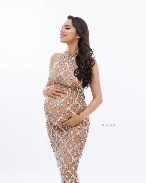 Maternity photography in Delhi: smiling expecting mother in a full-length crystal-embellished halter gown, eyes closed, hands on her bump