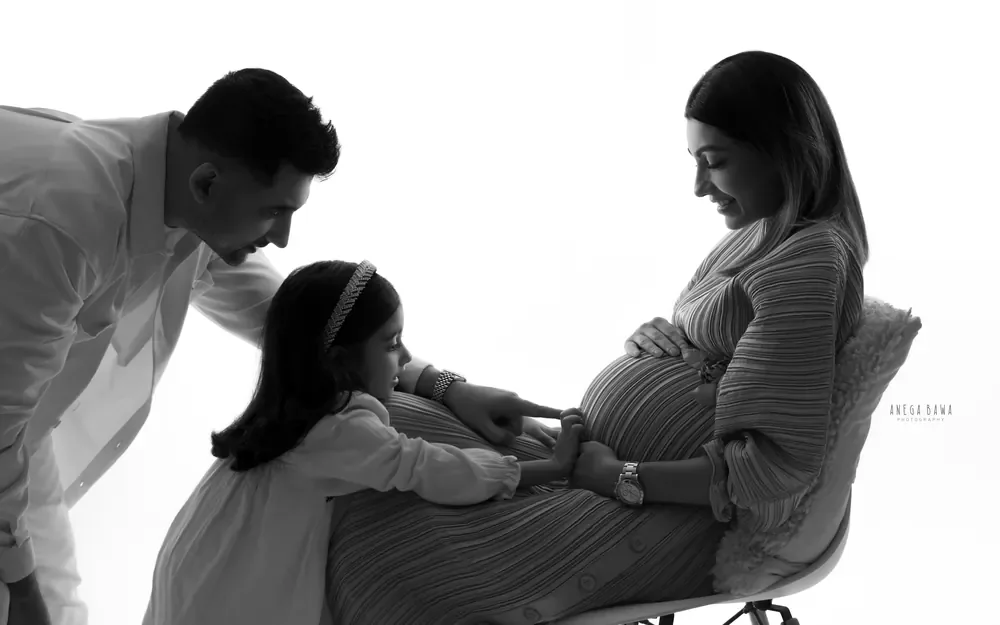 Maternity photography in Delhi: black and white portrait of a daughter touching her mother's bump as she sits on a white chair in a pleated