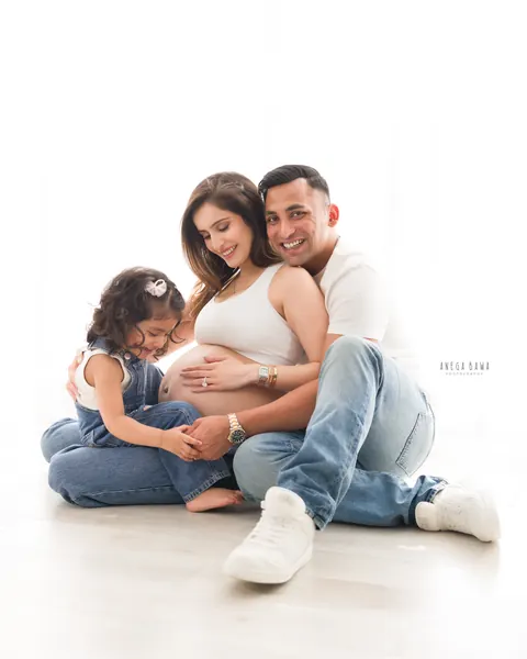 Maternity photography in Delhi: smiling parents with their toddler daughter seated on a white floor, all hands together on the bump, by