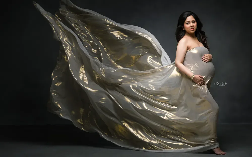 Maternity photography in Delhi: expecting mother in a strapless gold metallic drape with the fabric billowing behind her, dark studio