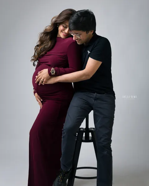 Maternity photography in Delhi: husband seated on a stool hugging his wife and holding her bump, she wears a burgundy gown, grey backdrop