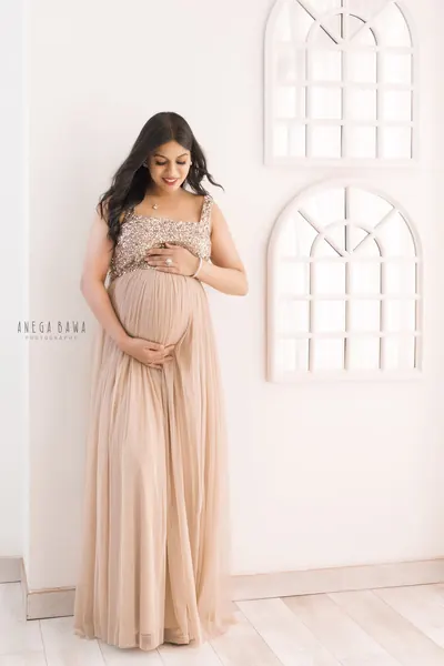 Smiling expecting mother in a blush sequin-bodice tulle gown with straps, looking down at her bump, standing against a white wall with two
