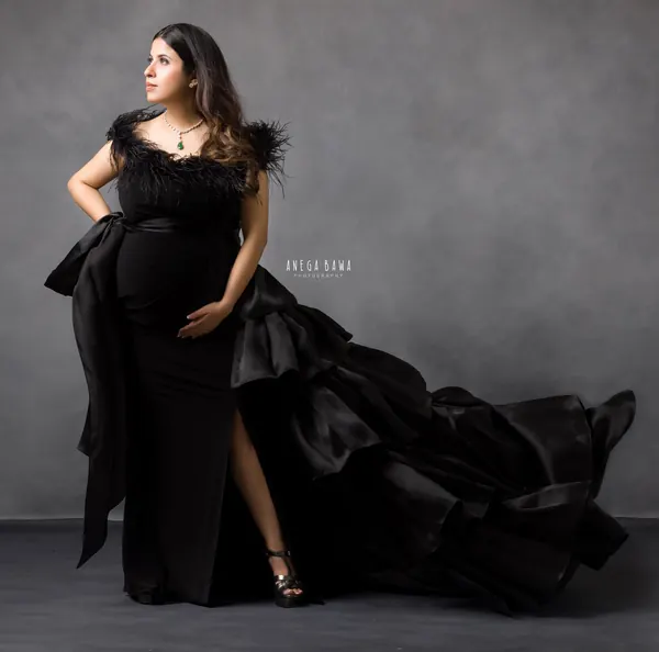 Expecting mother in a black fitted gown with a feather-trimmed neckline, black satin bow and a tiered black ruffle train swept out behind