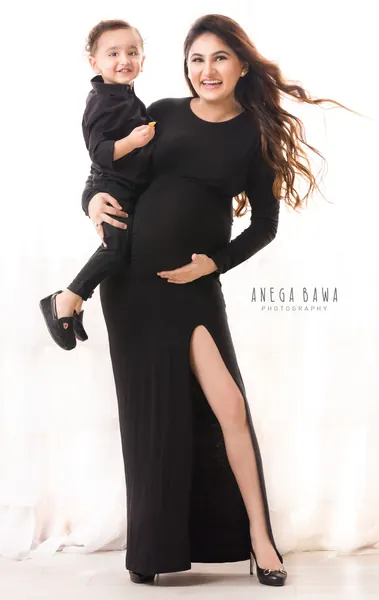 Smiling expecting mother in a black long-sleeve fitted gown with a high slit and heels carrying her toddler son in black on her hip, hair