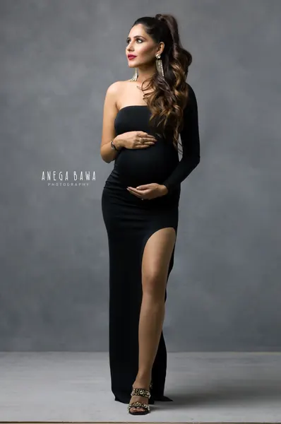 Expecting mother in a fitted black one-shoulder long-sleeve gown with a high thigh slit and embellished heels, hair in a high ponytail