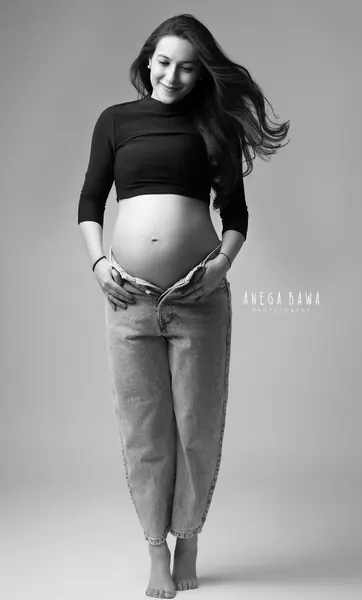 Black and white portrait of smiling expecting mother in a cropped black turtleneck and unzipped high-waisted jeans, bare bump, barefoot