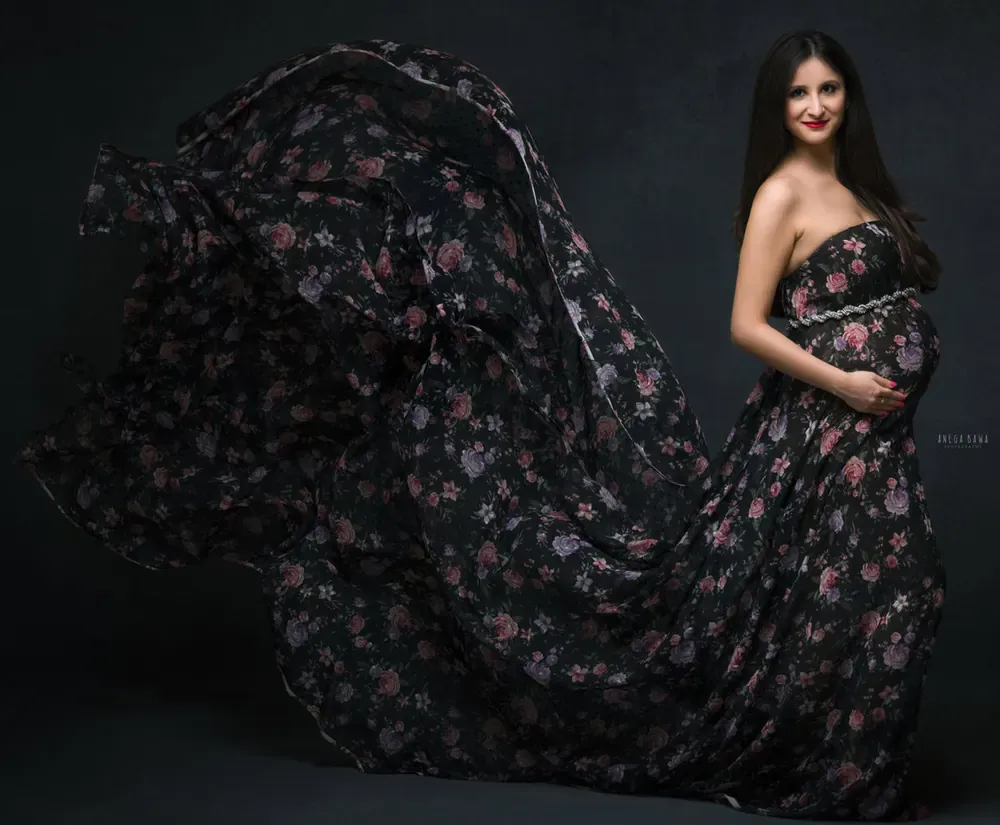 Expecting mother in a strapless dark floral gown with a very long sheer floral train flying out behind her, hand on her bump, dark backdrop.