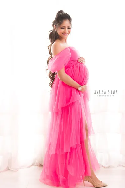 Smiling expecting mother in a bright pink tiered tulle off-shoulder gown with a high slit and nude heels, hands on her bump, hair in a high