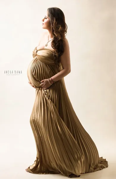Expecting mother in profile in a gold pleated satin strapless gown with a long flowing skirt, hands on her bump, looking up, warm cream
