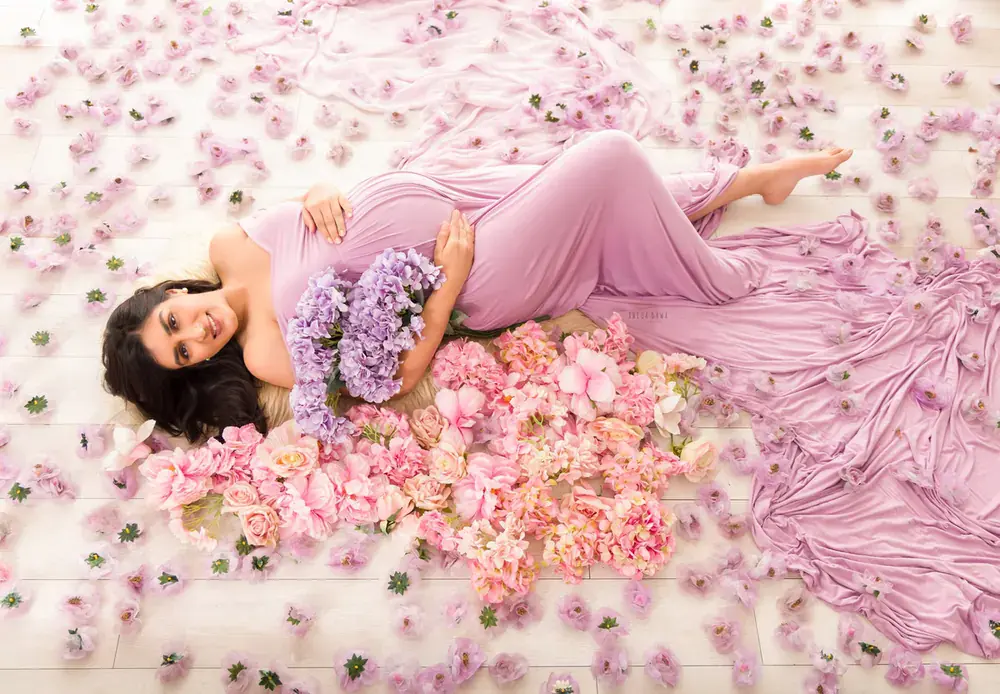 Overhead view of expecting mother lying in a lilac fitted gown among pink and lilac flowers and scattered petals on a white floor, holding