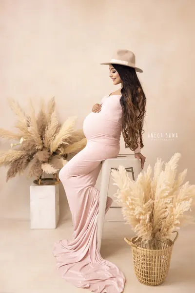 Expecting mother seated on a stool in a dusty pink fitted long-sleeve gown with a long train and a tan felt hat, surrounded by pampas