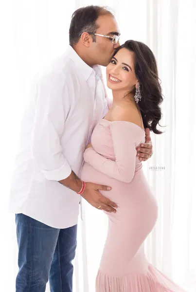 Smiling couple, husband in a white shirt and glasses kissing his wife's head and holding her bump, wife in a blush pink off-shoulder